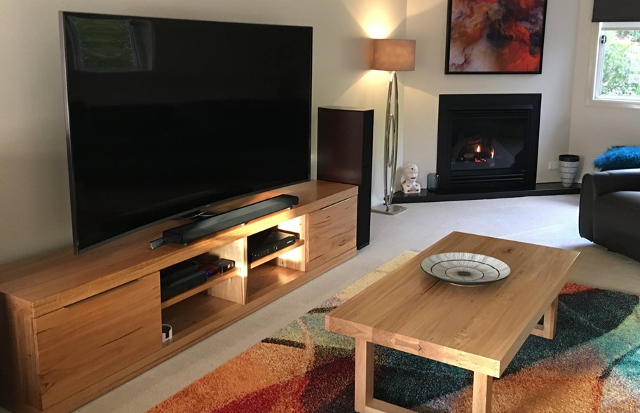 A cozy living room with a large flat-screen TV placed on a spacious wooden TV stand. The stand features multiple compartments for storage and a stylish design that complements the modern decor of the room, which includes a fireplace, a contemporary floor lamp, and a colorful rug.